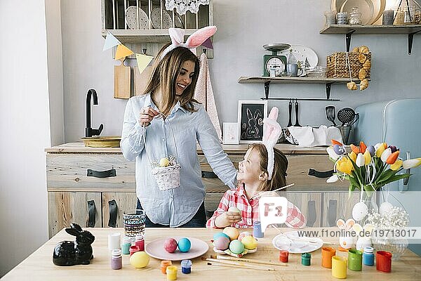 Glückliches Mädchen malt Eier Ostern in der Nähe von Mutter mit Korb