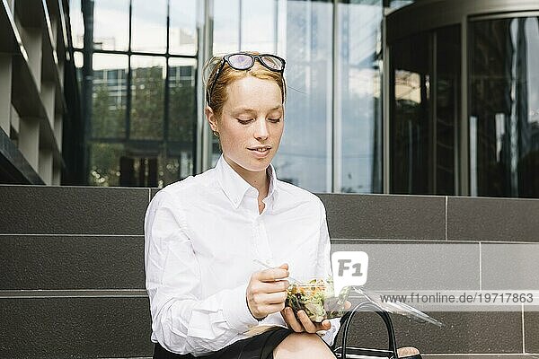 Junge Geschäftsfrau sitzt vor dem Büro und ißt ihre Lunchbox