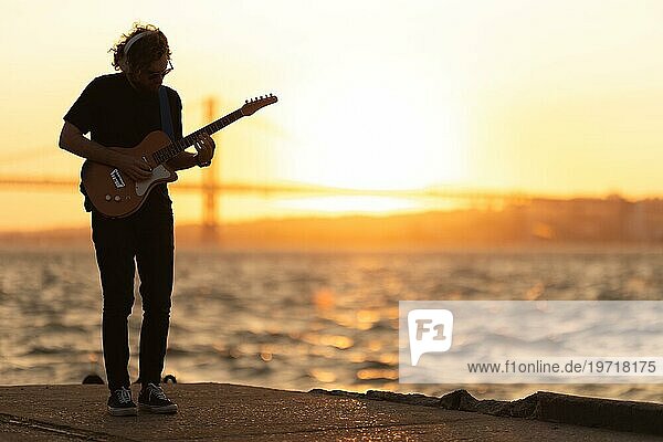 Ein Mann steht am Wasser und spielt bei Sonnenuntergang Gitarre. Mittlere Einstellung