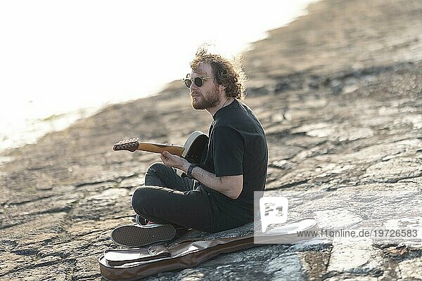Ein alleinstehender Hipster spielt Gitarre am Kai. Mittlere Einstellung