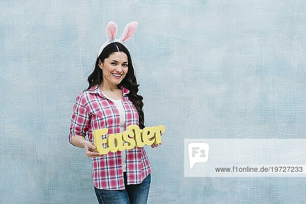 Lächelnde Frau mit Hasenohren zeigt Ostern Wort gegen blaün strukturierten Hintergrund