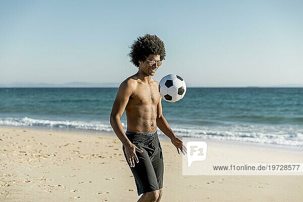 Lächelnder junger afroamerikanischer Mann spielt Fußball an der Seeküste