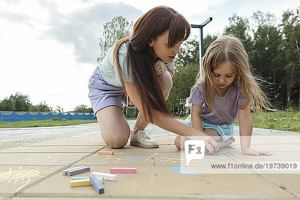 Woman teaching daughter to draw with chalk on tiled floor at park