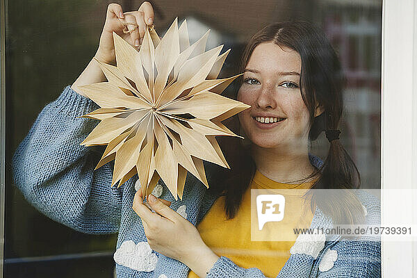 Smiling girl holding star shaped Christmas decoration seen through glass