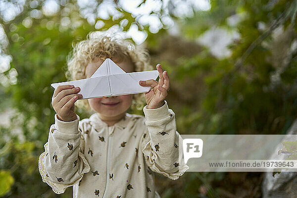 Happy girl holding paper boat over face in front of tree