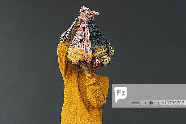 Woman covering face with fruits in mesh bags against black background