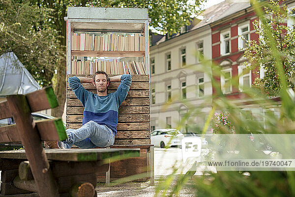 Man with eyes closed leaning on cabinet of books near plants in suburb