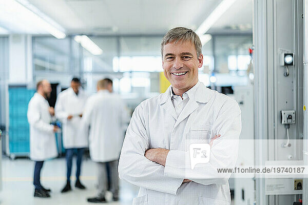 Technician in white lab coat standing in electronics factory with arms crossed