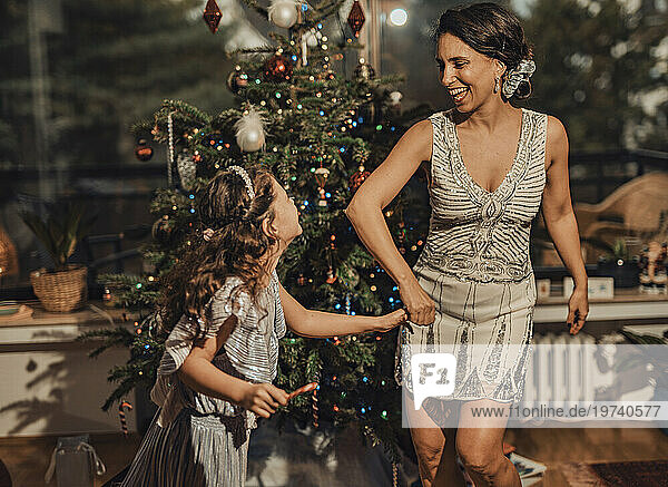 Happy girl holding hands with mother and dancing near Christmas tree at home