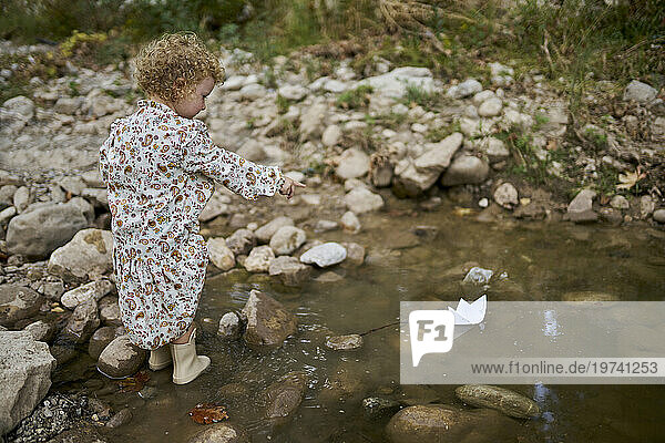 Cute girl playing and pointing on paper boat in water puddle near rocks