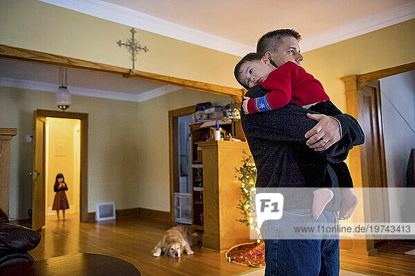 Father holds his young son in their home  with a young sister  dog and Christmas tree in the background; Lincoln  Nebraska  United States of America