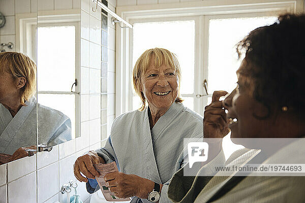 Happy blond woman talking to female friend while doing skincare in bathroom at home