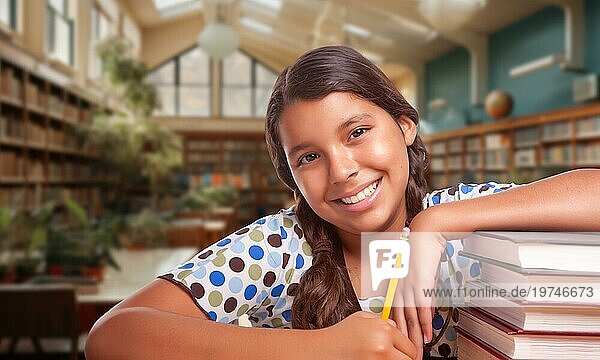 Glückliches junges hispanisches Mädchen mit einem Bleistift und einem Stapel Bücher bei den Hausaufgaben in einer Bibliothek