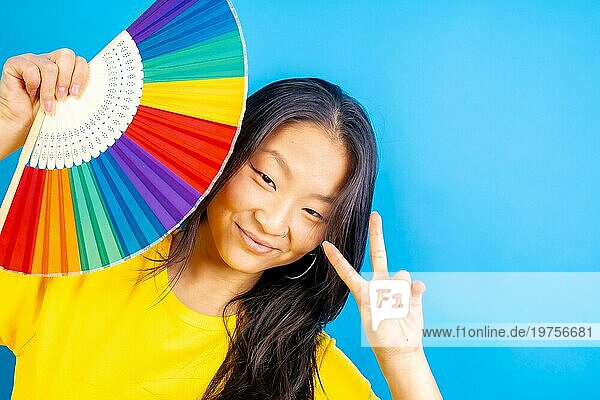 Studiofoto mit blauem Hintergrund von einer chinesischen Frau mit einem Regenbogen farbigen Fächer Geste Frieden