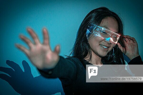 Studiofoto mit blauem Hintergrund mit Neonlichtern einer chinesischen Frau  die mit einer Augmented Reality Brille gestikuliert und Spaß hat