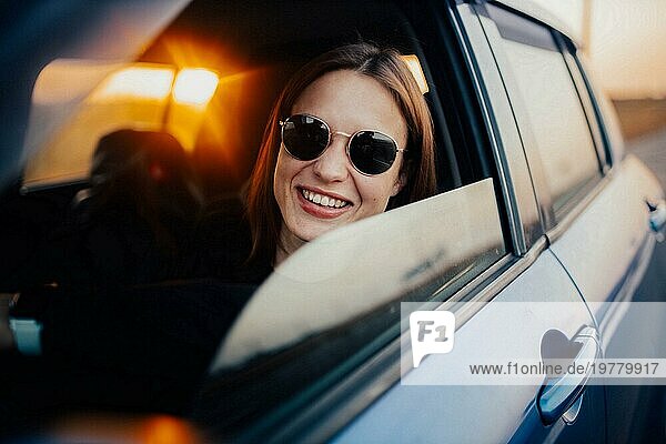 Glückliche Autofahrerin mit Sonnenbrille fährt ein Auto und lächelt bei Sonnenuntergang. Glückliche Reise Konzept