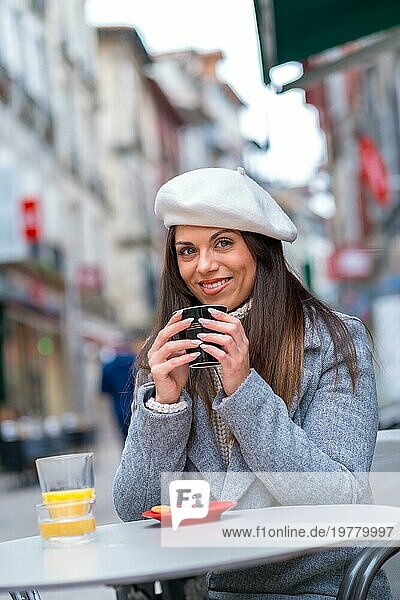 Vertikales Porträt einer Frau mit Baskenmütze  die in die Kamera lächelt  während sie im Freien Kaffee trinkt
