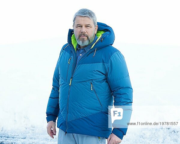 Mann  56 Jahre alt mit grauen Haaren und grauem Vollbart steht vor einer Schneefläche im Winter  seitlich zur Kamera  kritischer Blick  dicke Daunenjacke  weißer Hintergrund