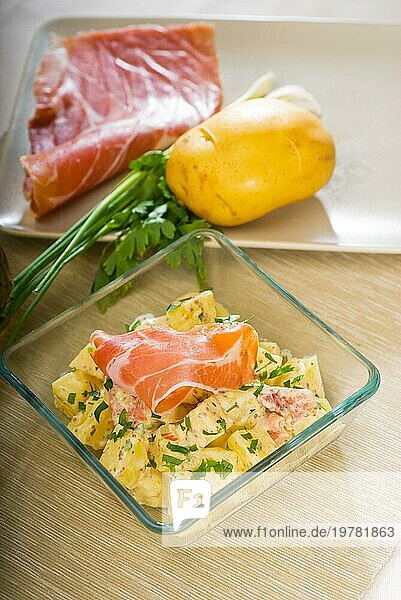 Frischer hausgemachter Parmaschinken und Kartoffelsalat  mit rohen Zutaten auf einem Tisch mit Schüsseln und Geschirr