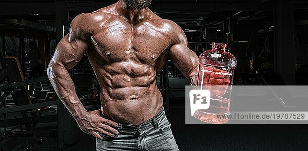 Muskulöser Mann steht in der Turnhalle mit einer großen Flasche Sportnahrung. Fitness und Bodybuilding Konzept. Gemischte Medien