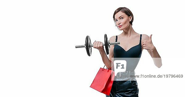 Schöne schlanke Frau posiert im Studio mit einer Hantel und Geschenktüten. Geschenk Konzept. Mitgliedschaft im Fitnessstudio. Ok Zeichen