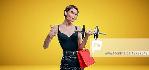 Schöne schlanke Frau posiert im Studio mit einer Hantel und Geschenktüten. Geschenk Konzept. Mitgliedschaft im Fitnessstudio. Ok Zeichen.
