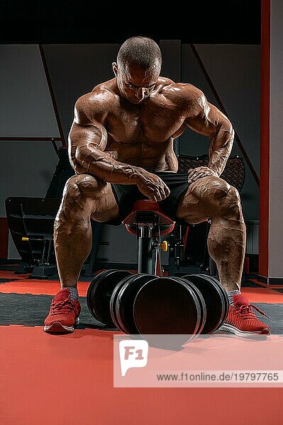 Muskulöser Mann sitzt auf einer Bank neben riesigen Hanteln. Bodybuilding und Kraftdreikampf Konzept.