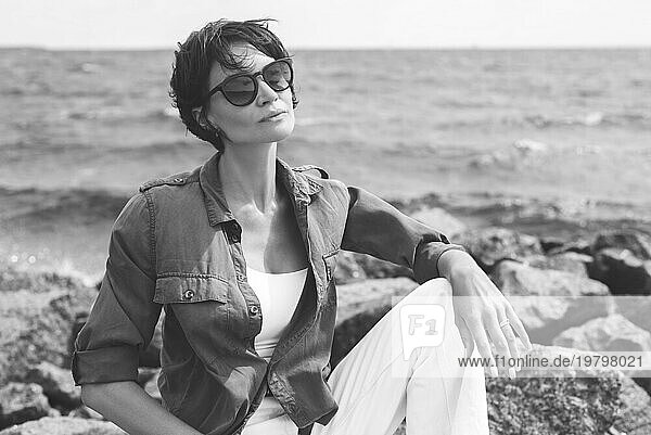 Porträt einer stilvollen Frau  die auf Steinen in der Bucht sitzt. Windiges Wetter. Fröhlichkeit  Freiheit und Sorglosigkeit. Tourismuskonzept
