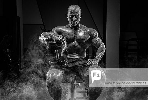 Muskulöser Mann sitzt auf einer Bank im Rauch mit Hanteln. Bodybuilding und Kraftdreikampf Konzept.