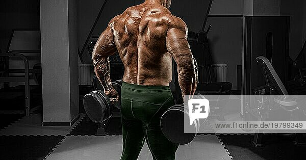 Kräftiger Bodybuilder posiert im Fitnessstudio mit Hanteln. Porträt ohne Namen. Rückansicht. Bodybuilding Konzept. Gemischte Medien