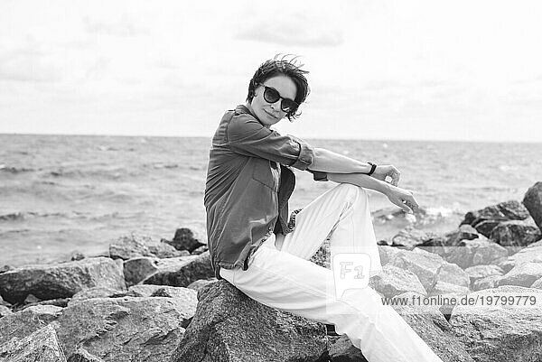 Porträt einer stilvollen Frau  die auf Steinen in der Bucht sitzt. Windiges Wetter. Fröhlichkeit  Freiheit und Sorglosigkeit. Tourismuskonzept