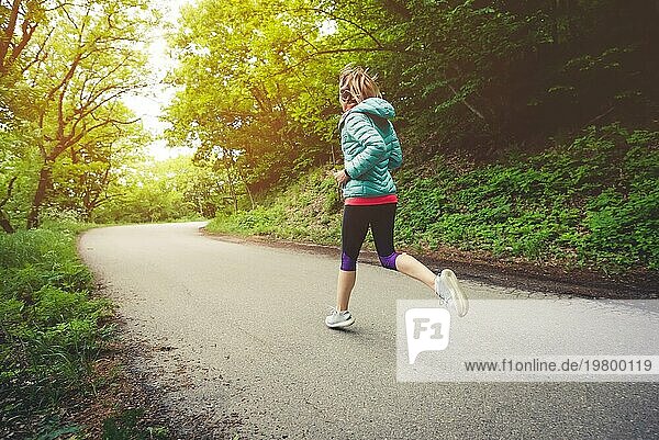 Junge Fitness blonde Frau mit Kopfhörern läuft am Morgen kaukasischen Waldweg in Sonne Licht. Seitenansicht von hinten