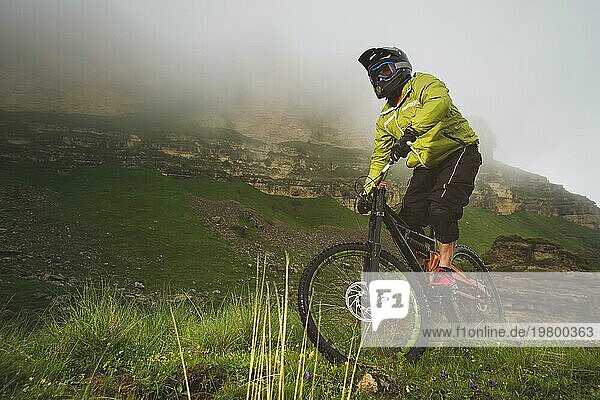 Ein erwachsener MTB Fahrer auf einem Mountainbike am Fuße einer Klippe  umgeben von grünem Gras. Tief hängende Wolken. Nordkaukasus. Russland