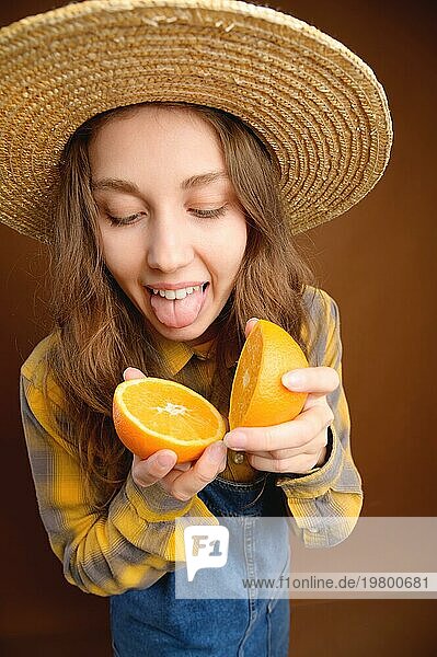 Studioporträt einer attraktiven kaukasischen jungen Frau mit Hut und Jeans Overall  die eine aufgeschnittene Orangenfrucht in den Händen hält und mit der Zunge versucht  sie zu kosten
