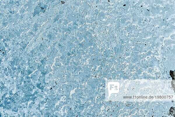 Nahaufnahme der Oberfläche eines Gletscherfragments mit einer Struktur aus Streifen und Blasen  die mit Sand und felsigem Gestein durchsetzt ist. Eisblaue Textur im Licht. Kleine Tiefenschärfe
