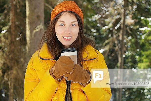 Attraktive positive kaukasische junge Frau in Winterkleidung mit Mütze  Handschuhen und Daunenjacke steht in einem winterlichen Nadelwald mit einem Pappbecher mit Kaffee oder Tee. Sie blickt in die Kamera. Raum kopieren