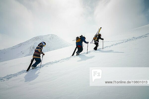 Drei befreundete Snowboarder und Skifahrer gehen mit Snowboard und Skiern in der Hand bergauf  um vor der Kulisse der schneebedeckten Berge des alpinen Skigebiets Backcountry oder Freeride zu fahren. Skitourenkonzept mit einer Gruppe von Menschen