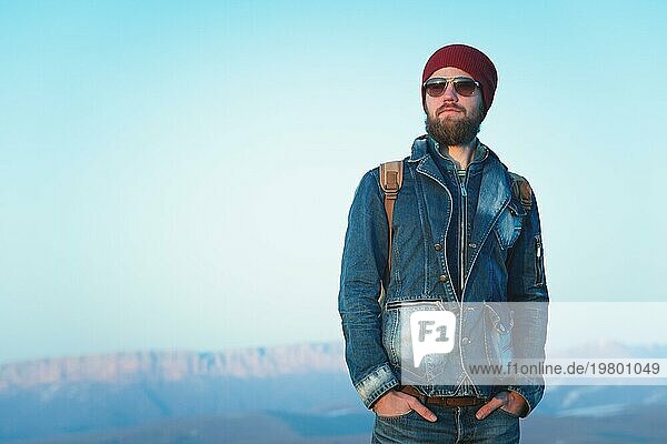 Modeporträt eines bärtigen jungen Mannes mit Sonnenbrille  Rucksack und Hut auf einem Hintergrund mit Copyspase in den Bergen bei Sonnenuntergang  Ein selbstbewusster Mann mit einem Bart