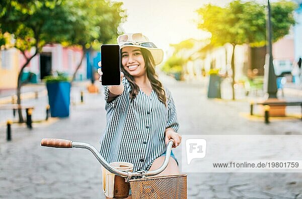 Schönes Mädchen mit Hut auf dem Fahrrad zeigt Handybildschirm auf der Straße. Lächelnde Tourist auf dem Fahrrad zeigt Handybildschirm auf der Straße