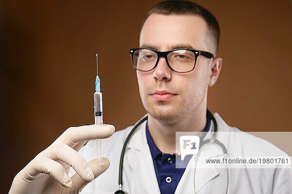 Junger kaukasischer Arzt  der sich darauf vorbereitet  eine Impfung mit einer Spritze durchzuführen. Studioaufnahme auf einem braunen Hintergrund. Lassen Sie Luft aus der Spritze vor der Injektion