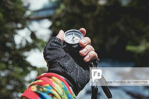 Eine behandschuhte Hand hält einen Magnetkompass vor dem Hintergrund eines Nadelwaldes und eines Gebirgsflusses. Das Konzept der Outdoor Navigation