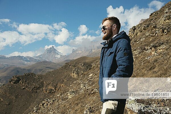 Hipster Reisender in Daunenjacke und Sonnenbrille steht an einem Berghang vor der Kulisse epischer Felsen und lächelt. Das Konzept  das Glück in den Bergen zu finden