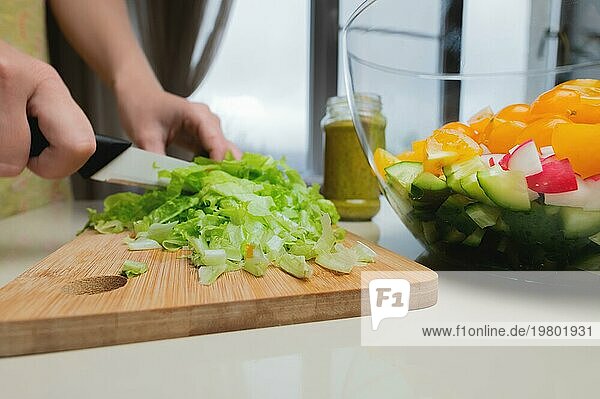 Weibliche Hände schneiden Salatblätter mit einem Messer vor dem Hintergrund einer Schüssel mit gehacktem Gemüse. Vegetarisches Essen Vorbereitung