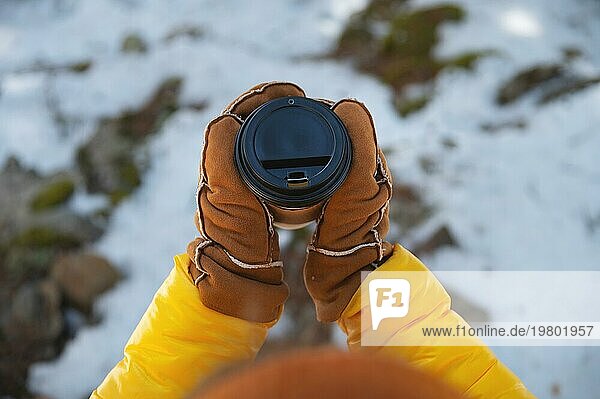 Ein Pappbecher mit einem schwarzen Deckel in den Händen in Handschuhen  in der Natur vor einem Hintergrund von Schnee
