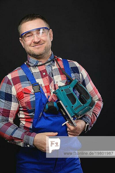 Porträt eines kaukasischen männlichen Mechanikers in Arbeitsuniform mit einer elektrischen Stichsäge um den Hals. Atelierporträt eines handwerklichen Geschäftsmannes