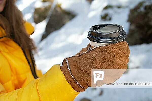 Nahaufnahme eines Pappbechers mit schwarzem Deckel und einem heißen Getränk in den Händen einer jungen Frau mit Handschuhen vor einem Hintergrund aus Natur und Schnee