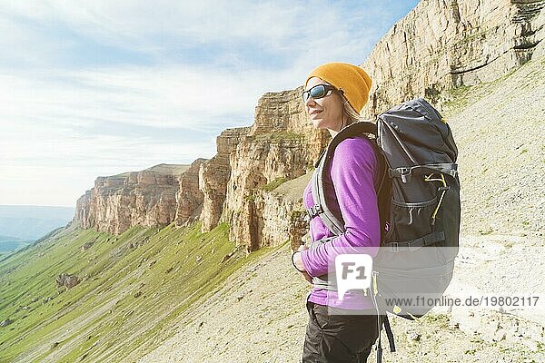 Lächelnde Reisende mit einem gelben Hut und einer Sonnenbrille steht am Fuße eines epischen Felsens mit einem Rucksack daneben und schaut weg