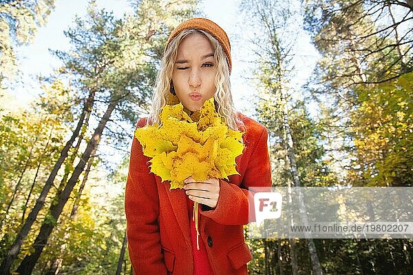 Attraktive weiße kaukasische Mädchen zwinkern in roten Polo und orange Hut ist mit einem Blumenstrauß von Blättern in ihren Händen vor dem Hintergrund der Herbstwald verwirrt. Das Konzept der Herstellung einer Entscheidung über etwas im Herbst im Park Wald