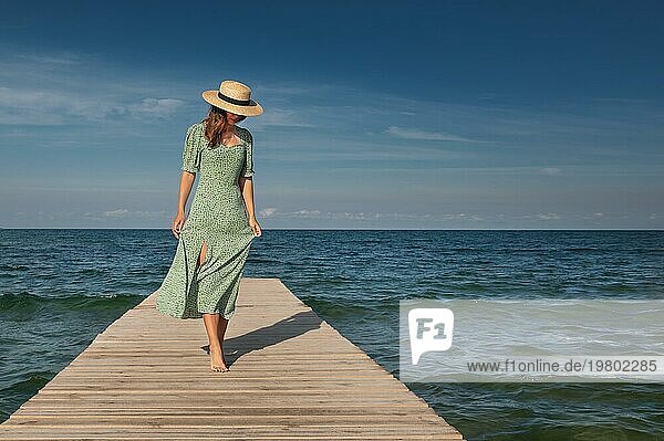 Eine attraktive junge kaukasische Frau in einem grünen Kleid und einem Strohhut geht an einem sonnigen Tag vor der Kulisse des Meeres leicht auf einem Holzsteg spazieren. Ruhe und Spaziergänge am Meer