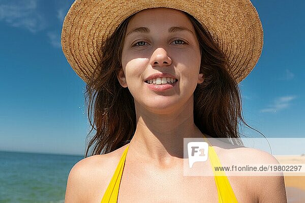Nahaufnahme einer attraktiven kaukasischen jungen Frau mit Strohhut und gelbem Badeanzug  die gegen den blaün Himmel und das Meer an einem sonnigen Tag lächelt